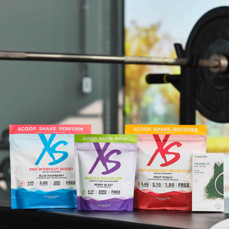 The Peak Performance Stack, featuring XS Pre-Workout Boost, XS Muscle Multiplier, XS Post-Workout Recovery and Nutrilite Vitamin B Dual Action. Products arranged on a table in a home gym setting with weights in the background.-en-US-m-01.jpg