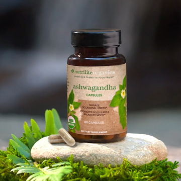 Nutrilite Organics Ashwagandha bottle on a rock in front of a waterfall. Two capsules arranged to the side.