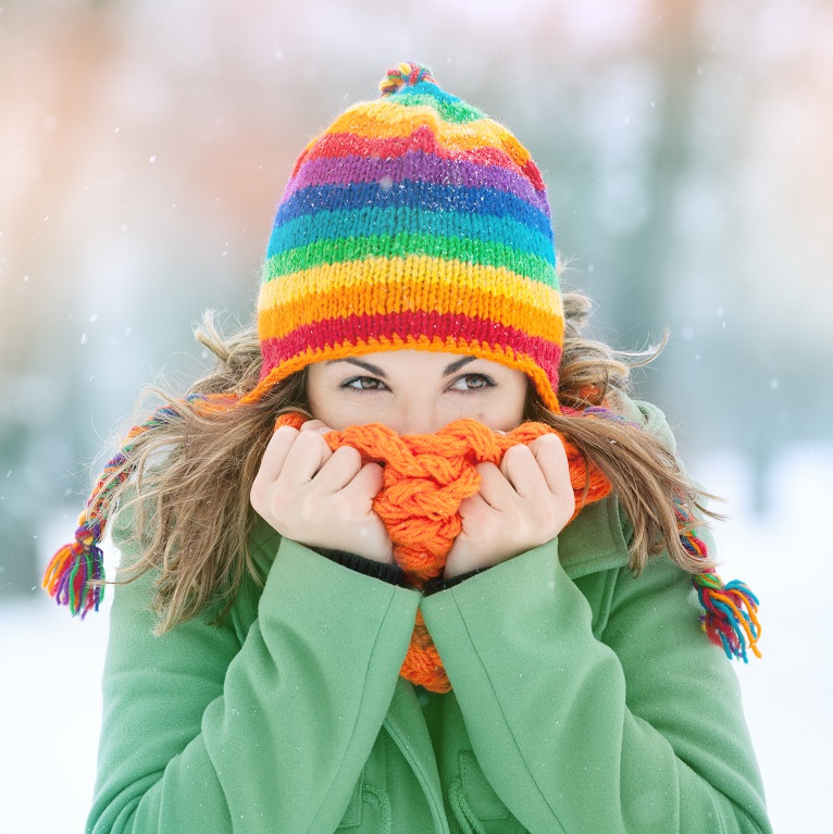 Una mujer con un abrigo verde y un gorro de lana de colores sostiene una bufanda naranja sobre su boca y nariz tratando de mantenerse abrigada en invierno.