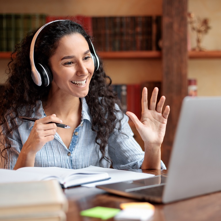 A smiling woman wearing headphones waves at the participants of her virtual class on a laptop. Bookshelves are visible in the background.