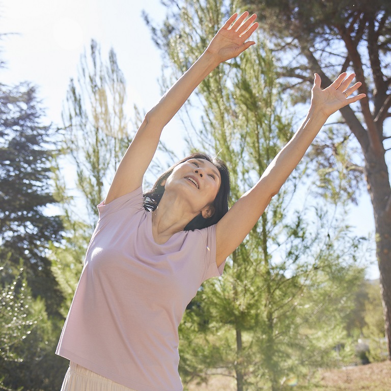 A woman stretches, leaning with her arms over her head, as she practices tai chi outside in the sun with trees in the background.