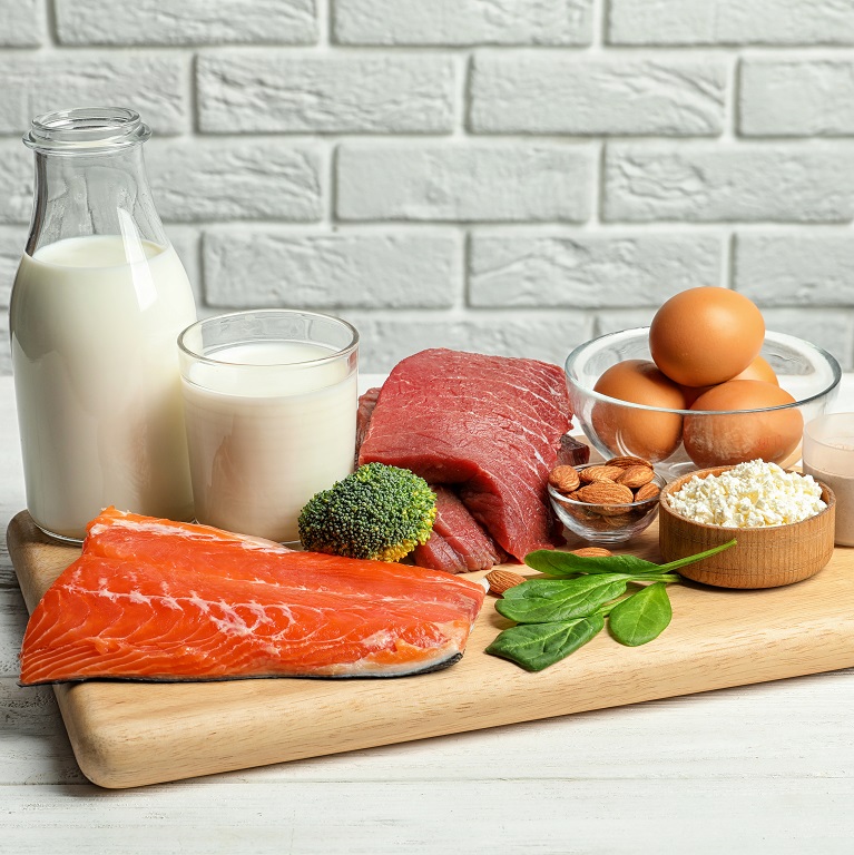A wooden cutting board displays sources of protein, including milk, salmon, beef, eggs, almonds, broccoli and spinach.