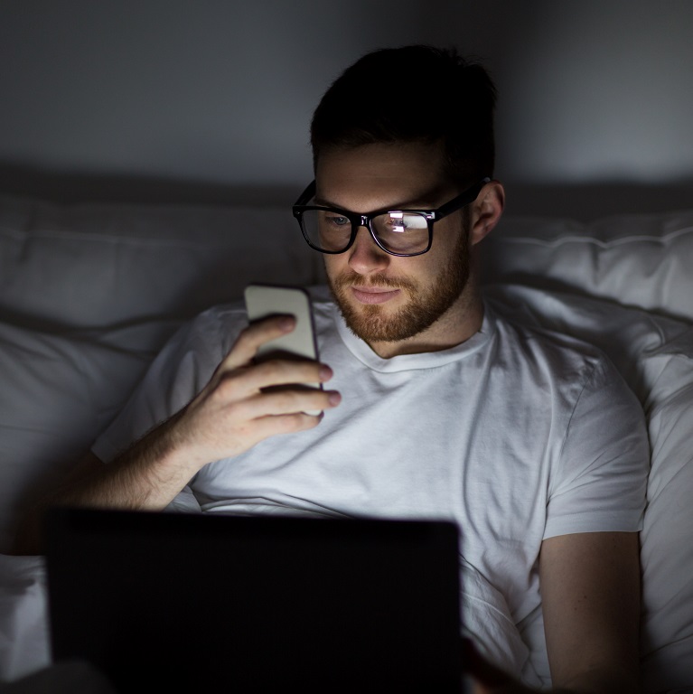 A man in bed stares at his phone with an open laptop: Two things that will not help you fall asleep.
