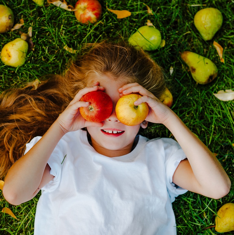 Vista superior de una niña acostada en el césped rodeada de manzanas y peras. Sostiene una manzana sobre cada ojo.