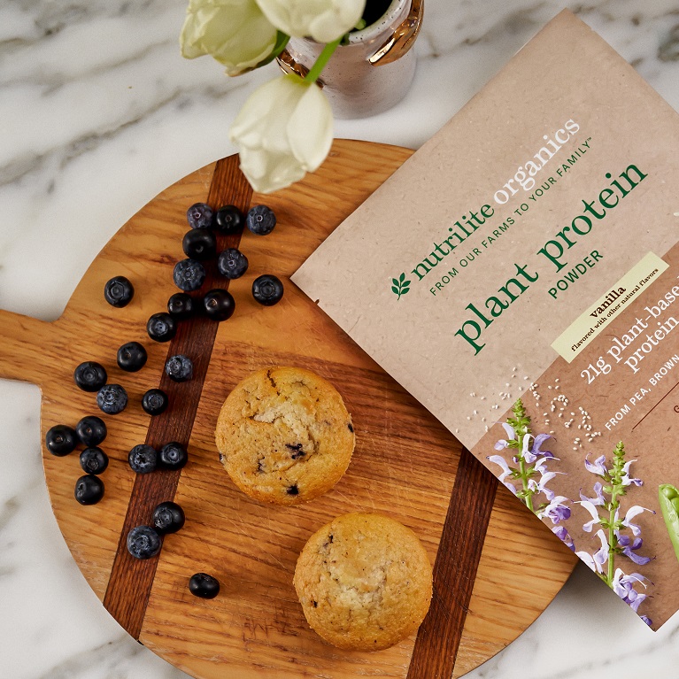 A package of Nutrilite Organics Vanilla Plant Protein Powder lies on a wooden cutting board next to blueberry muffins, blueberries, flowers and a whisk.