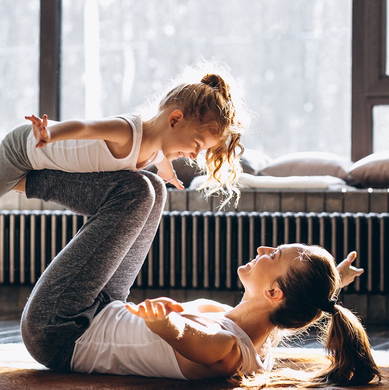 A smiling woman in yoga clothes lies on the floor lifting her small daughter on her legs.