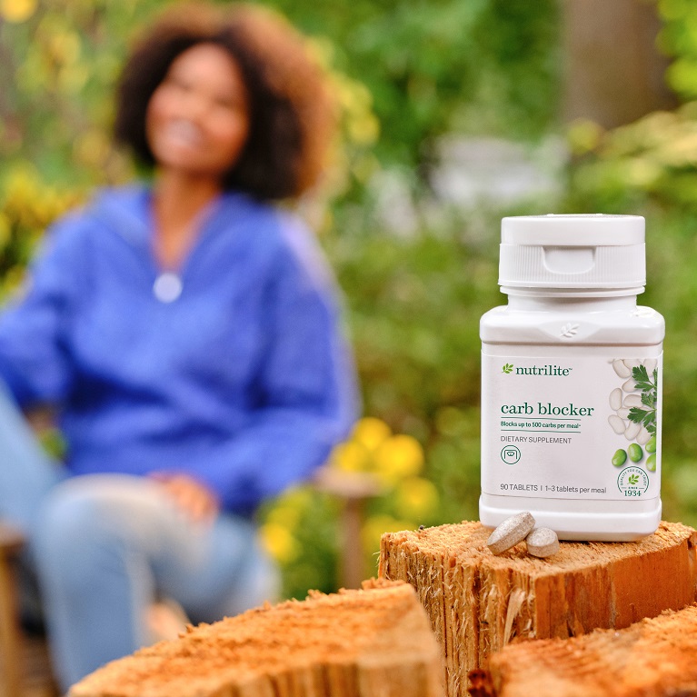 A bottle of Nutrilite Carb Blocker is in the foreground while a smiling woman drinking wine is in the background.