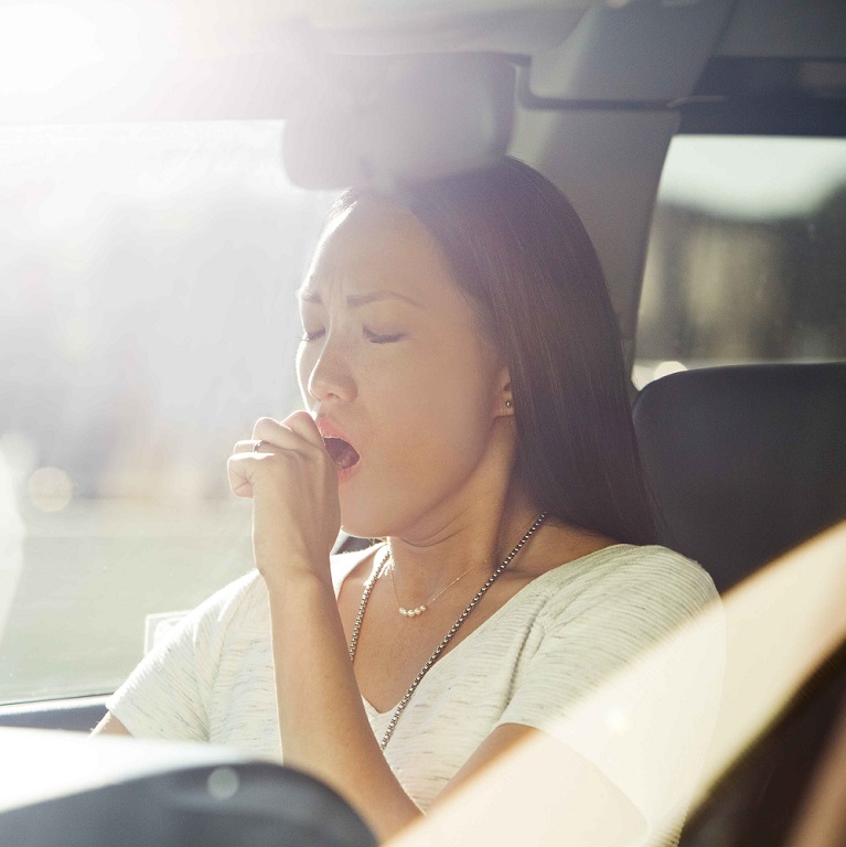 A woman sitting in the front seat of a car holds her hand in front of her mouth during a yawn.
