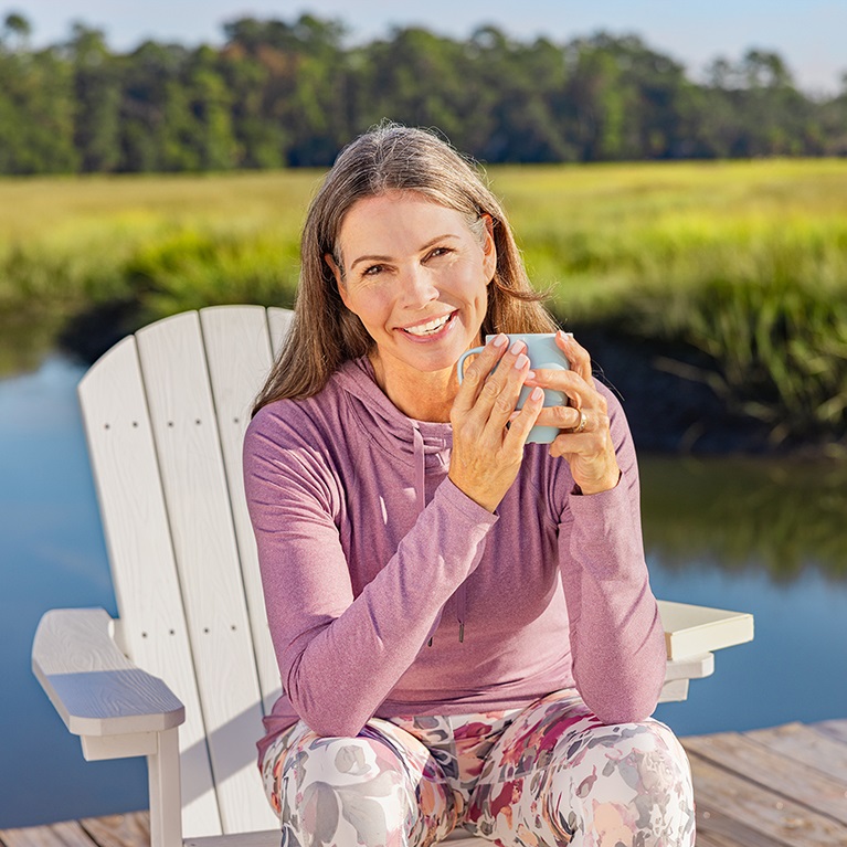 A smiling woman holding a coffee or tea mug sits in an Adirondak chair on a dock. Water, a green field and trees are in the background.