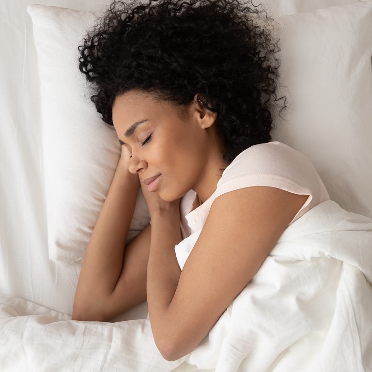Una mujer de cabello rizado oscuro durmiendo de lado en una cama con sábanas blancas. Con la cabeza apoyada sobre sus manos.