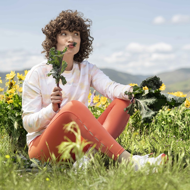 Una mujer sentada cerca de un campo de flores amarillas come felizmente col rizada verde y frondosa.