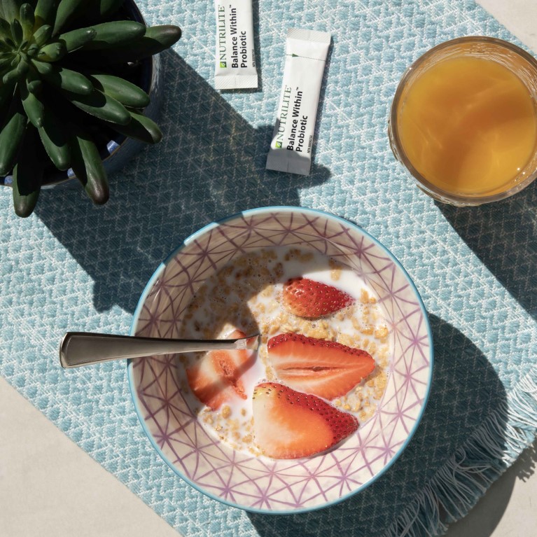Several items sit on a blue placemat in the outdoor sun, including a succulent plant, cereal with milk and strawberries, orange juice and Nutrilite Balance Within Probiotics.