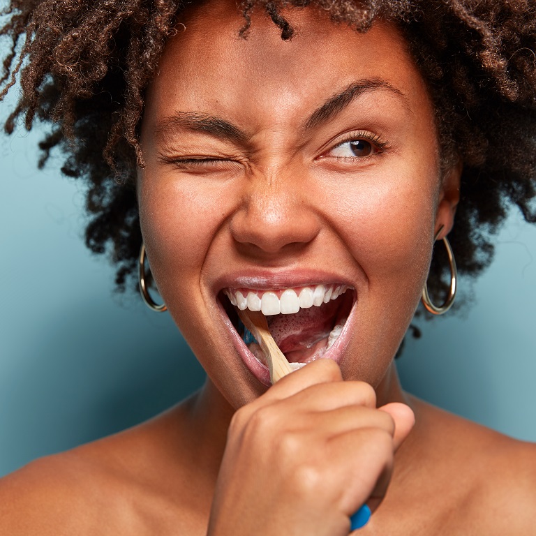Una mujer sonriente frente a un fondo azul se cepilla los dientes. Su ojo derecho está cerrado mientras su ceja izquierda está levantada de una manera peculiar.