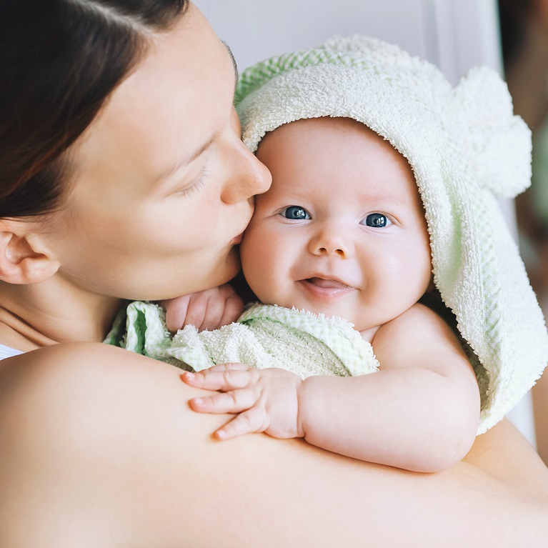 A woman holds a smiling baby fresh from the bath. The baby is wrapped in a green-hooded towel.