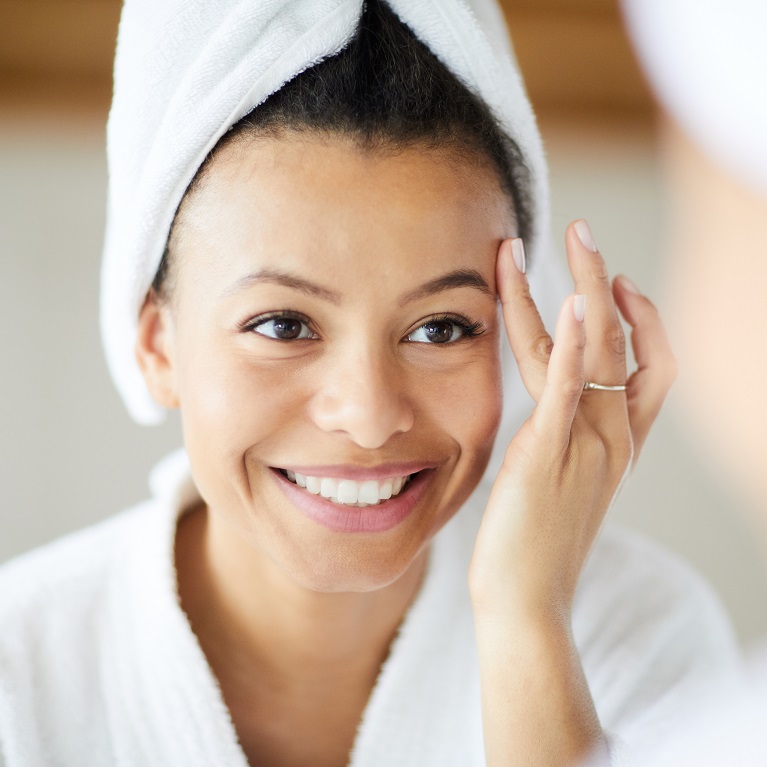 Una mujer sonriendo en una bata de baño blanca con el pelo envuelto en una toalla aplicándose un producto para el cuidado de la piel Artistry en la frente con el dedo anular.
