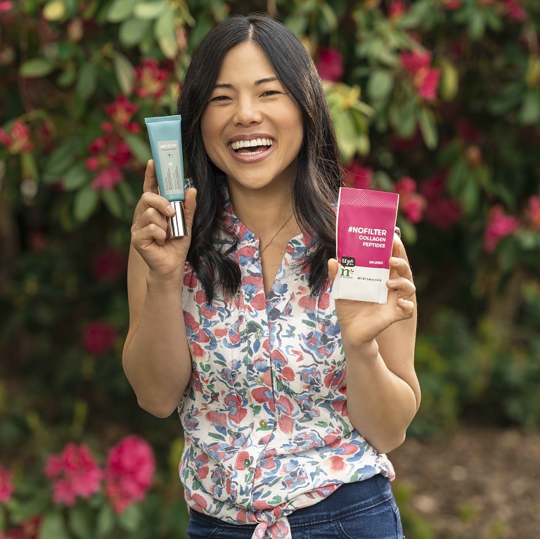 Una mujer sonriendo sostiene un tubo de Crema de día de reactivación renovadora Artistry Skin Nutrition y un paquete de Péptidos de colágeno #sinfiltro n* by Nutrilite.