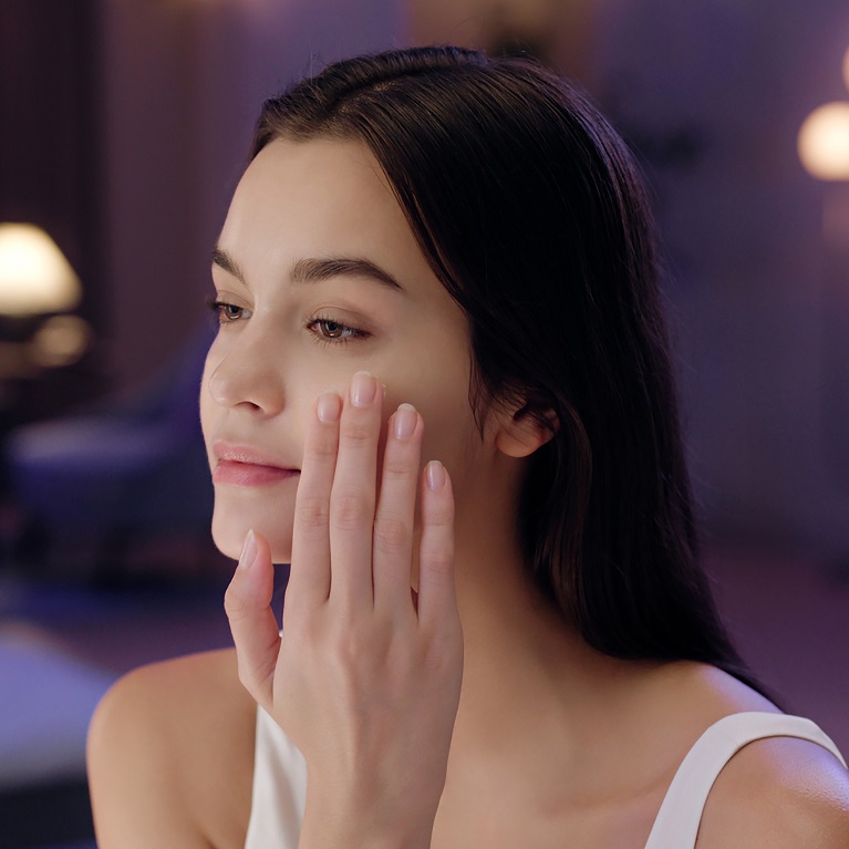 A woman in a dimly-lit bedroom applies an Artistry Skin Nutrition Sleeping mask to her face.