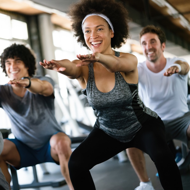 Three smiling people do squats in a group fitness class at a gym.