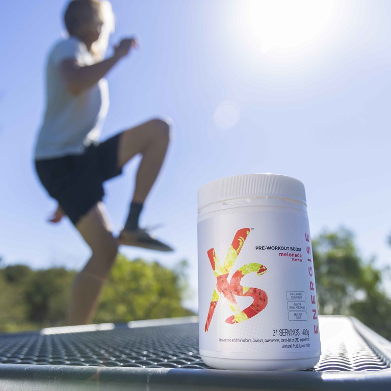 A container of XS Pre-Workout Boost sits on a bench outside. A person is seen exercising in the background.